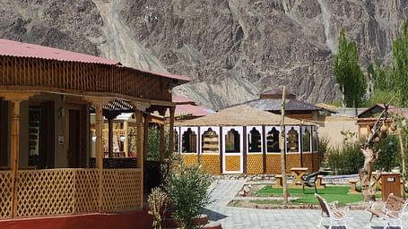 Outdoor seating area and traditional wooden structures with mountain backdrop, featuring a garden path and part of a circular blue-tiled pool.