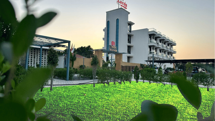 A modern hotel building is seen beyond a green lawn, with plants in the foreground and a clear sky above - Lords SKD Resort, Salangpur