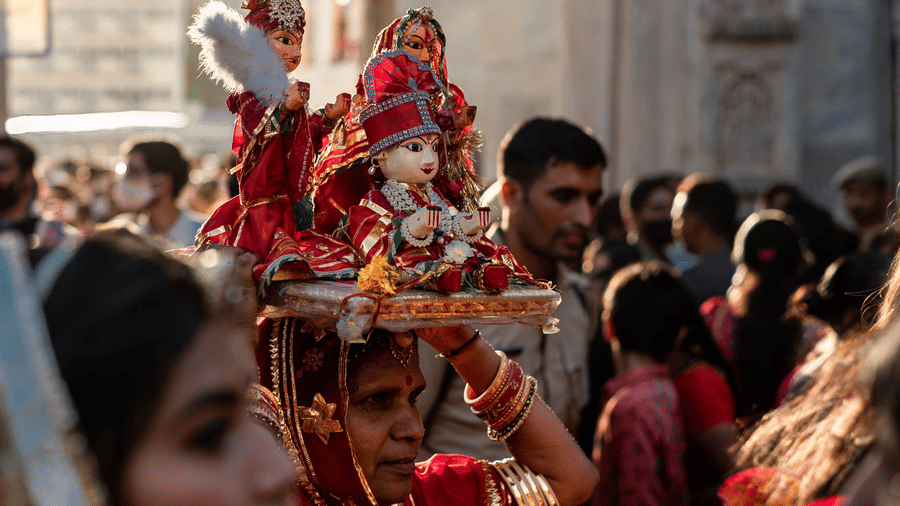 A woman dressed in Rajasthani traditional outfit holding an idol of goddess Gauri in a crowded procession with other devotees.