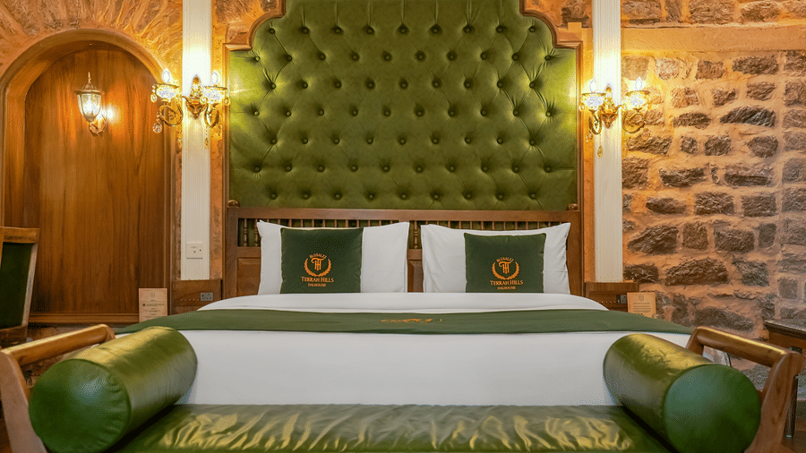 The premium room at BluSalzz Terrah Hills Resort, Dalhousie, featuring a double bed with white linens, a tall tufted headboard, and a upholstered bench at the foot of the bed, all set against a stone wall. 