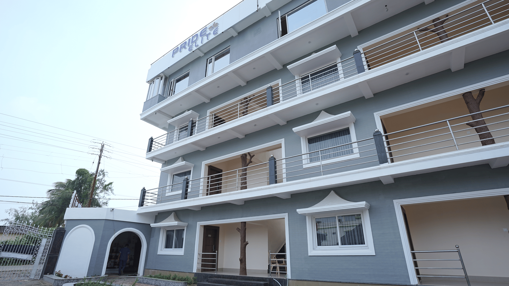 Exterior of Pride Elite Mandarmani with the Pride Elite signage, grey and white multi-storey facade, balconies with decorative tree sculptures, and a gated entrance.