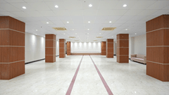 Empty banquet hall at Pride Elite Mandarmani with wood-panelled pillars on both sides, a raised stage in the background, and a pink sofa near the entrance.
