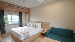 Guest room at Pride Elite Mandarmani with a bed with grey upholstered headboard, a teal sofa, wooden coffee table, bedside phone, and a window overlooking the pool.