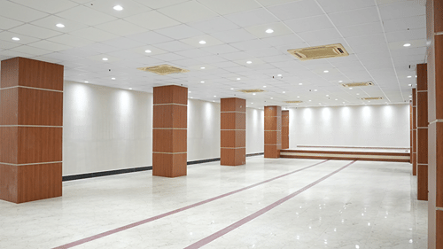 Angled view of an empty banquet hall at Pride Elite Mandarmani with a row of wood-panelled pillars leading toward a raised stage across marble flooring.