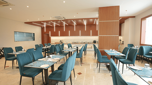 Restaurant at Pride Elite Mandarmani with teal chairs at multiple laid tables around a central wood-clad pillar, marble flooring, and a buffet counter.