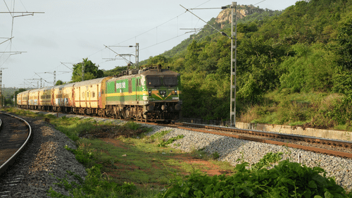 A long passenger train with green and yellow coaches travels along a curved track through a lush green landscape with a large hill in the background.