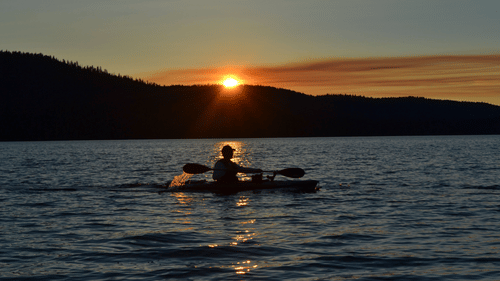 A solitary person riding a kayak with a paddle in hands while the sun is seen setting over a hill in the background.