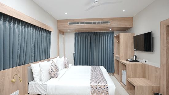 Guest room at Pride Elite Mandarmani with a bed with patterned cushions facing a window, an open wardrobe, wall-mounted TV, and a coffee station.