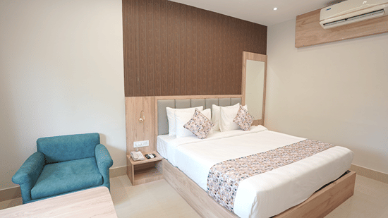 Guest room at Pride Elite Mandarmani with a wooden platform bed, grey headboard, teal armchair, full-length mirror, and a wall-mounted air conditioner.