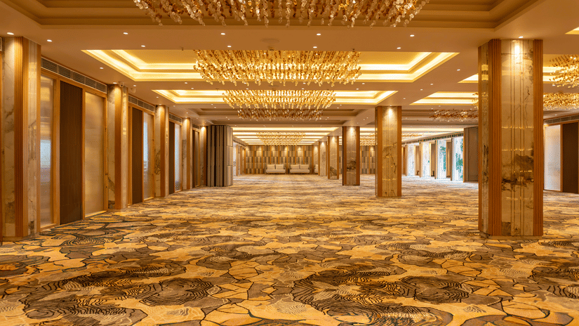 The grand banquet hall at Pride Elite Himmatnagar features elegant golden lighting and spacious patterned carpeting.