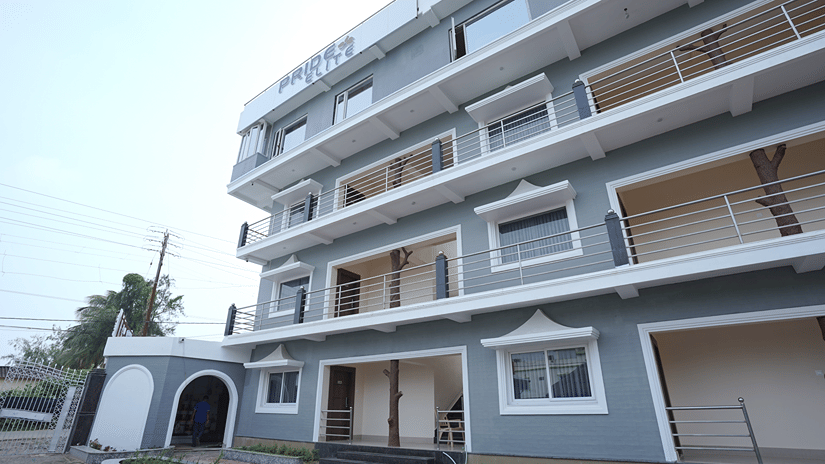 Exterior of Pride Elite Mandarmani with the Pride Elite signage, grey and white multi-storey facade, balconies with decorative tree sculptures, and a gated entrance.