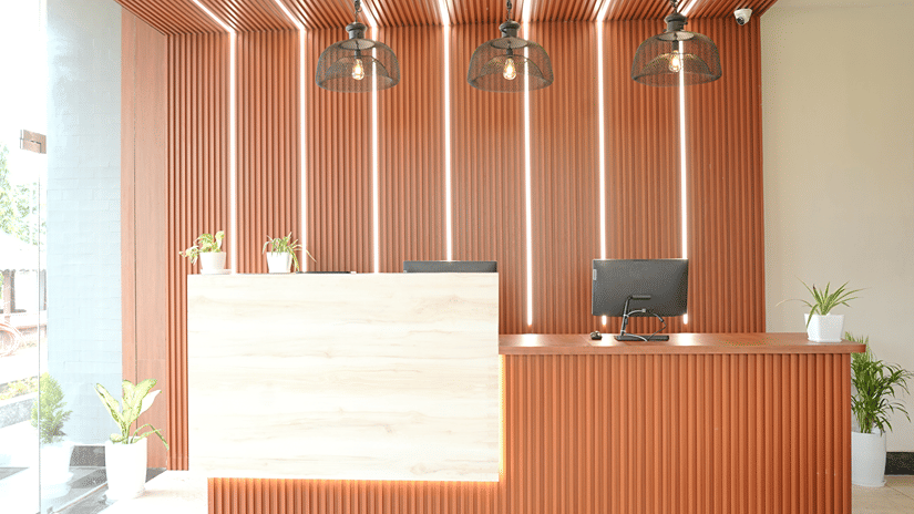 Close-up of the reception desk at Pride Elite Mandarmani with a wooden counter, monitor, 3 cage pendant lights, ribbed wood panelling, and potted plants.
