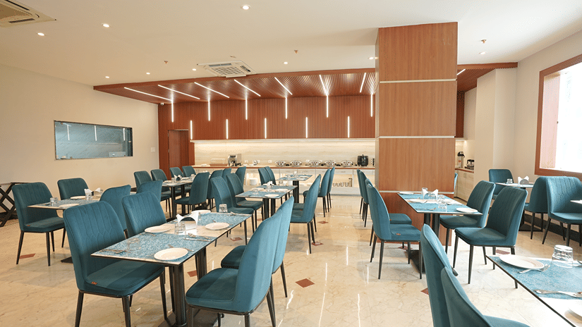 Restaurant at Pride Elite Mandarmani with teal chairs at multiple laid tables around a central wood-clad pillar, marble flooring, and a buffet counter.