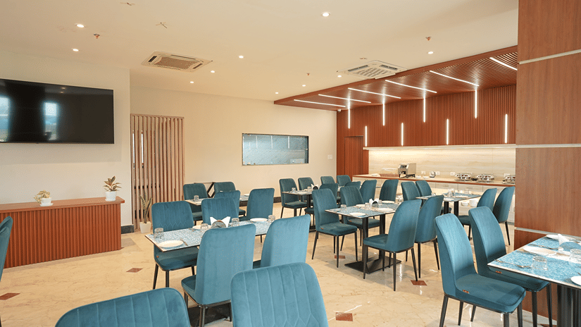 Restaurant at Pride Elite Mandarmani with teal dining chairs, a wall-mounted television, potted plants, and a wood-slat ceiling above the buffet counter.