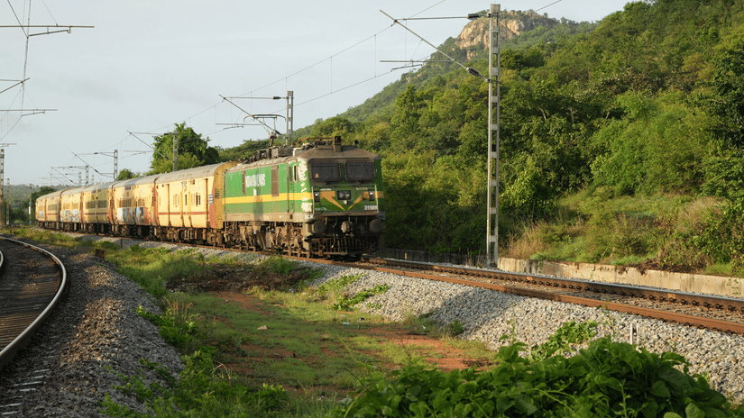 A long passenger train with green and yellow coaches travels along a curved track through a lush green landscape with a large hill in the background.