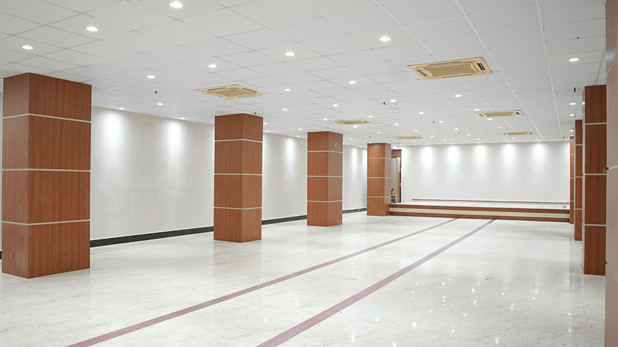 Empty banquet hall at Pride Elite Mandarmani with closely spaced wood-panelled pillars, a recessed ceiling with cassette air conditioners, and a raised stage.