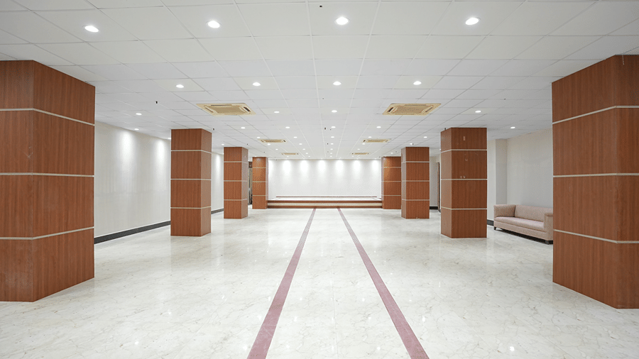 Empty banquet hall at Pride Elite Mandarmani with wood-panelled pillars on both sides, a raised stage in the background, and a pink sofa near the entrance.