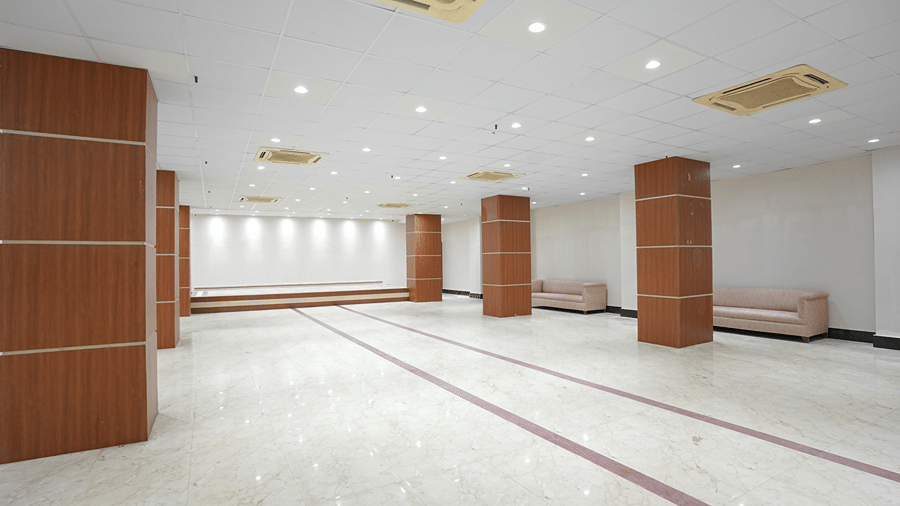 Empty banquet hall at Pride Elite Mandarmani with polished marble flooring, wood-panelled pillars, a raised stage, and pink sofas along the wall.
