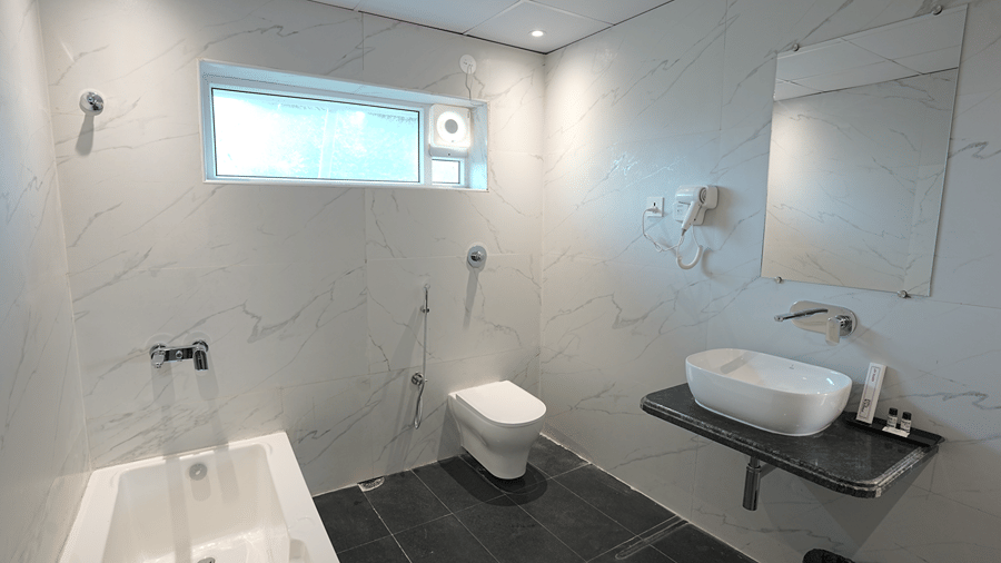 Bathroom at Pride Elite Mandarmani with a bathtub, a wall-mounted toilet, a vessel basin on a dark countertop with mirror above, and black floor tiles.