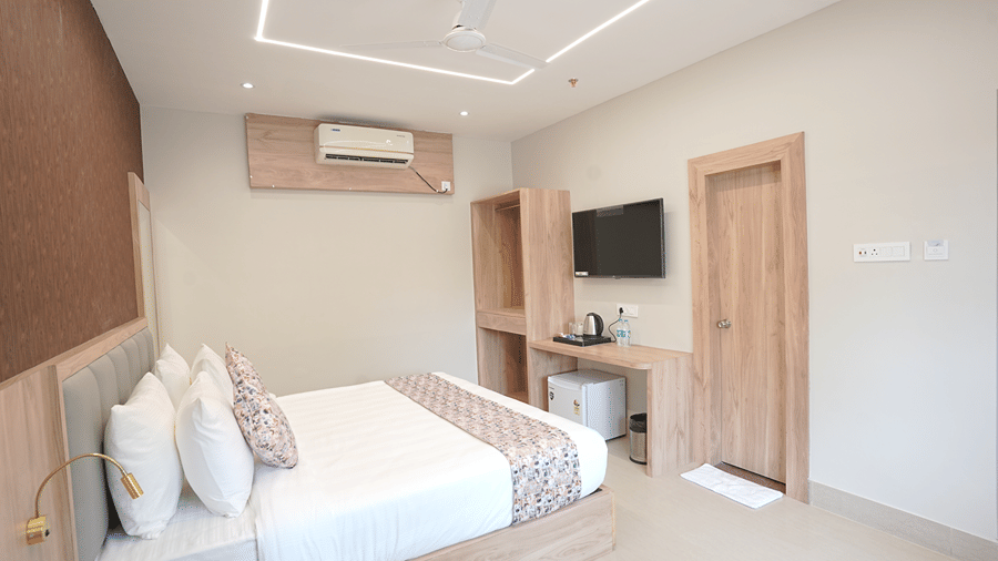 Guest room at Pride Elite Mandarmani with a bed with grey headboard, ceiling fan, wall-mounted TV, open wardrobe, mini fridge, and wooden entry door.