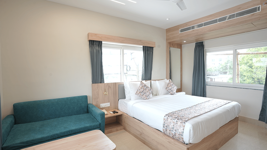 Corner guest room at Pride Elite Mandarmani with 2 curtained windows, a teal sofa, wooden coffee table, and a bed with patterned cushions.