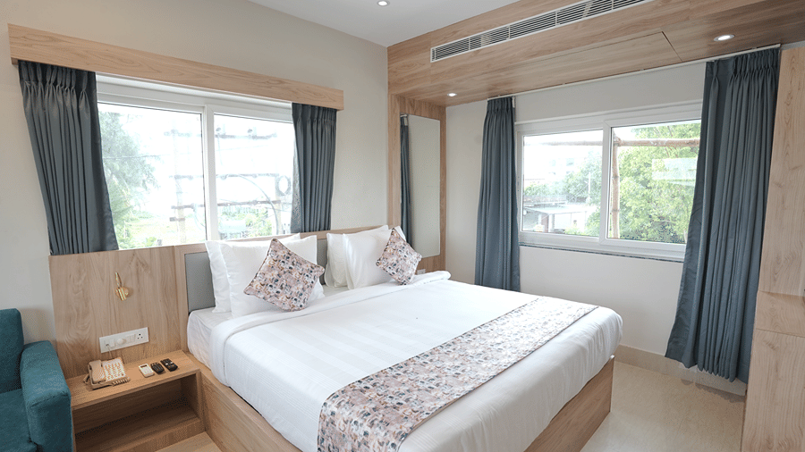 Guest room at Pride Elite Mandarmani with a bed with patterned cushions, 2 corner curtained windows, a full-length mirror, and a bedside table with a phone.