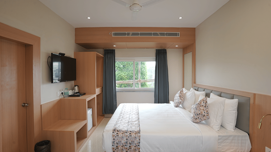 Guest room at Pride Elite Mandarmani with a bed with patterned runner and grey headboard, wall-mounted TV, wooden storage unit, and a window with grey curtains.