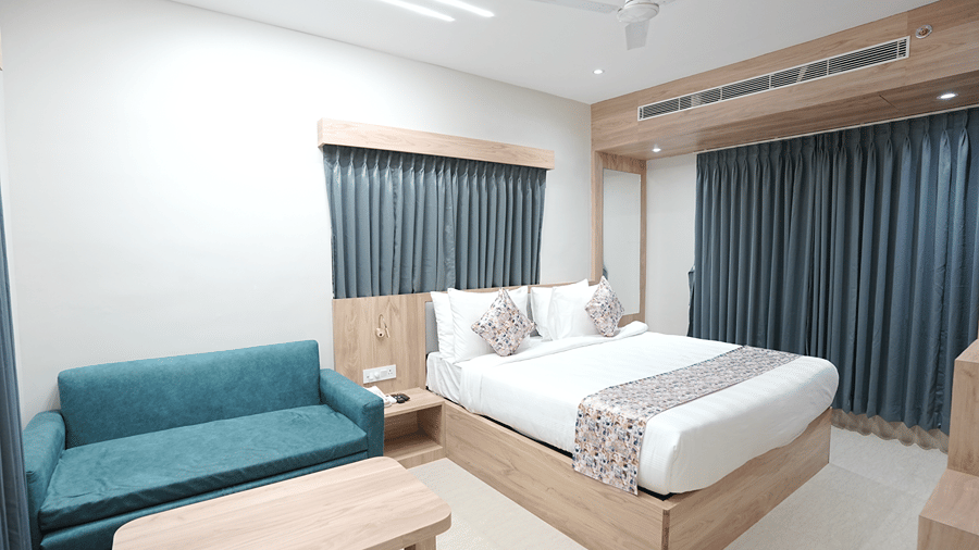 Guest room at Pride Elite Mandarmani with a teal sofa, wooden coffee table, a bed with patterned cushions, and floor-length curtains on 2 windows.