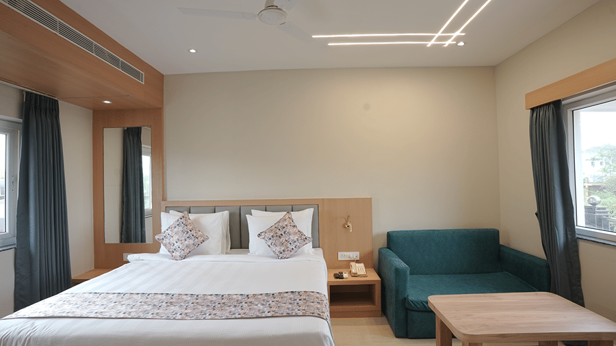 Guest room at Pride Elite Mandarmani with a bed, grey headboard, bedside telephone, teal sofa, wooden coffee table, full-length mirror, and 2 curtained windows.
