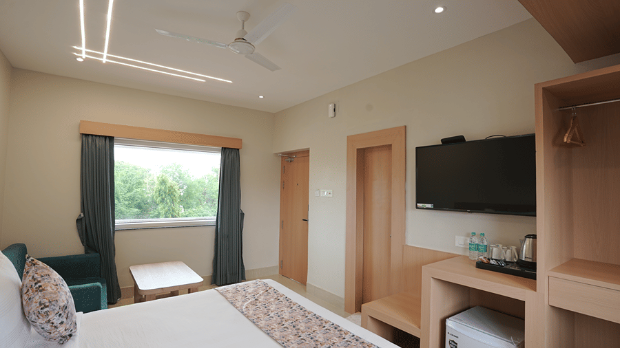 Guest room at Pride Elite Mandarmani with a bed, teal armchair by a window with a garden view, wall-mounted TV, wooden wardrobe, tea and coffee station, and mini fridge.