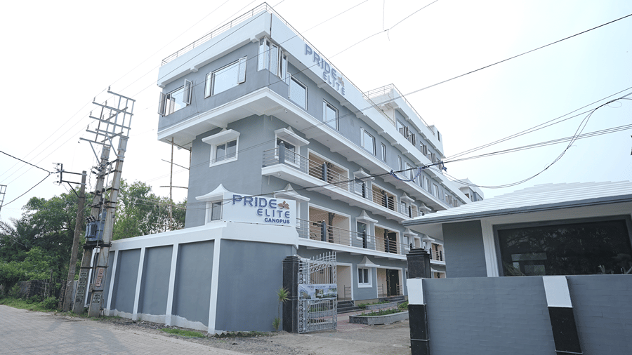 Exterior of Pride Elite Mandarmani with the Pride Elite Canopus signage, grey and white multi-storey facade, balconies, a gated entrance, and boundary wall.
