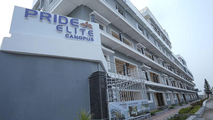 Exterior of Pride Elite Mandarmani with the Pride Elite Canopus signboard, grey and white multi-storey facade, wrought-iron gate, and a we are now open banner.