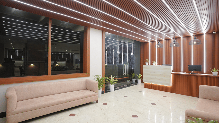 Lobby at Pride Elite Mandarmani with a wide angle of the reception desk, ribbed wooden ceiling with linear lighting, pendant lamps, pink sofas, potted plants, and a glass entrance.
