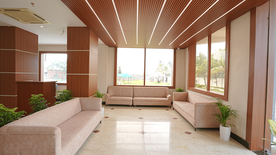 Lounge area at Pride Elite Mandarmani with 3 pink sofas, wood-clad columns, ribbed wooden ceiling with linear lighting, and windows overlooking palm trees and gazebos.