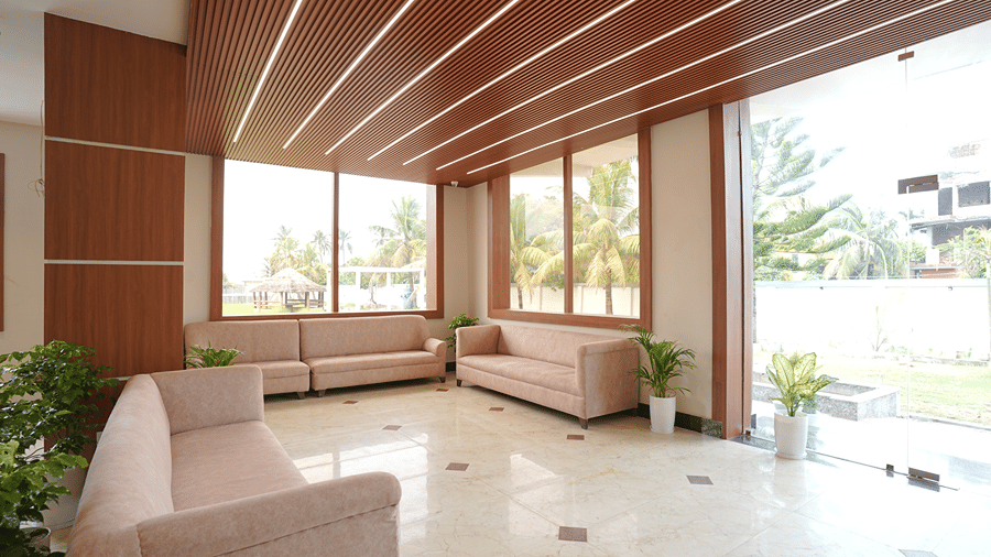 Lounge seating area at Pride Elite Mandarmani with pink sofas, potted plants, ribbed wooden ceiling with linear lighting, and large windows overlooking palm trees and lawn.