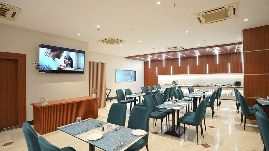 Restaurant at Pride Elite Mandarmani with several laid tables and teal chairs, a wall-mounted TV, and a clear view of the buffet counter with chafing dishes.