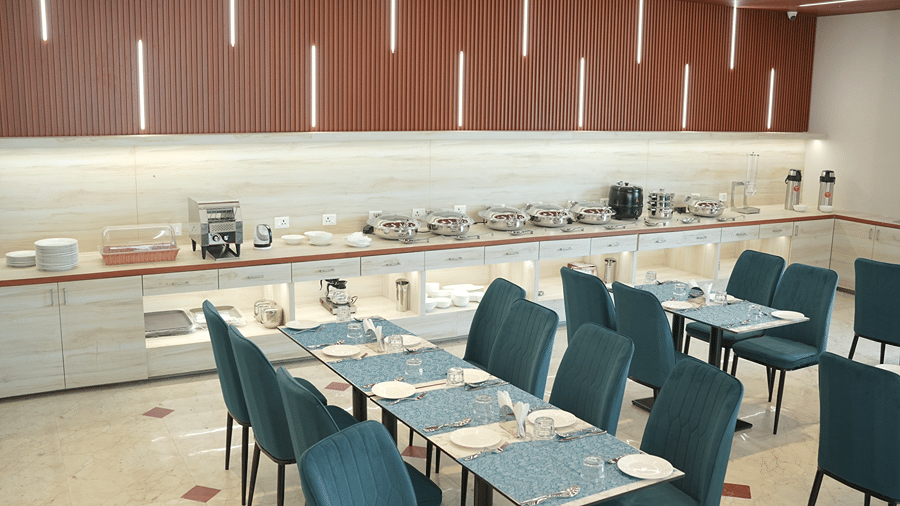Restaurant at Pride Elite Mandarmani with laid dining tables and teal chairs in the foreground, and a buffet counter with chafing dishes behind.