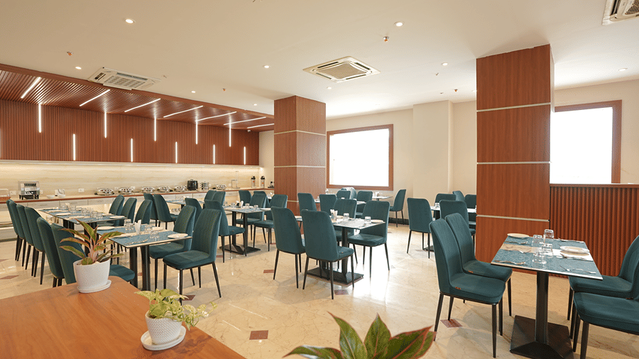 Restaurant at Pride Elite Mandarmani with potted plants in the foreground, teal chairs at dining tables, wood pillars, and large windows letting in daylight.