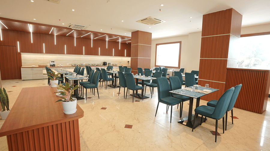Restaurant interior at Pride Elite Mandarmani seen from a wooden counter with potted plants, showing teal chairs at dining tables and large windows.