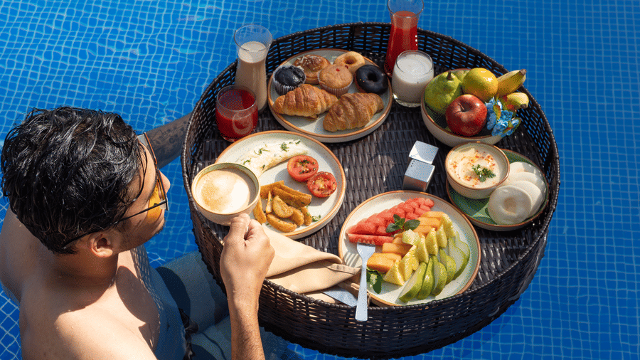 person holding a cup of coffee near the floating tray holding breakfast items and drinks in a blue swimming pool at The Golden Tusk, Jim Corbett