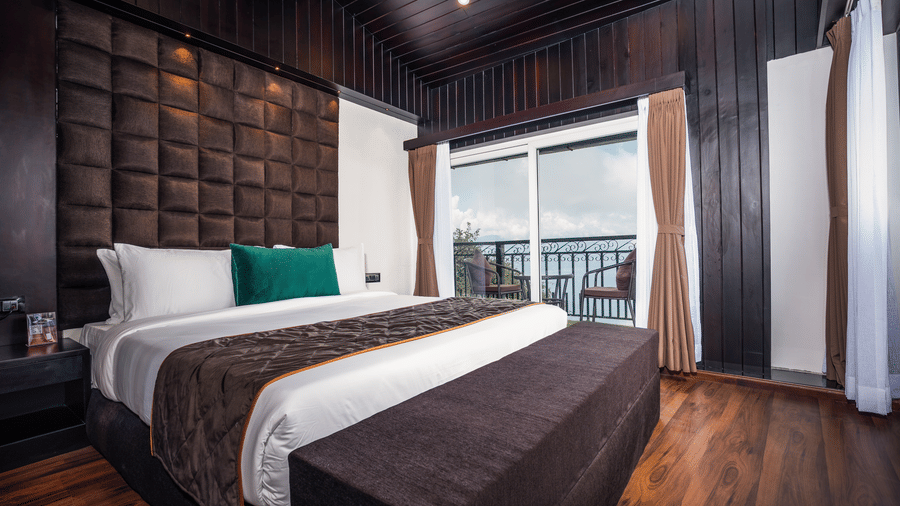 Bedroom at The West Gate Posada By Summit, Darjeeling, showing a bed, padded headboard, side table, wooden floor, and sliding door to a balcony.