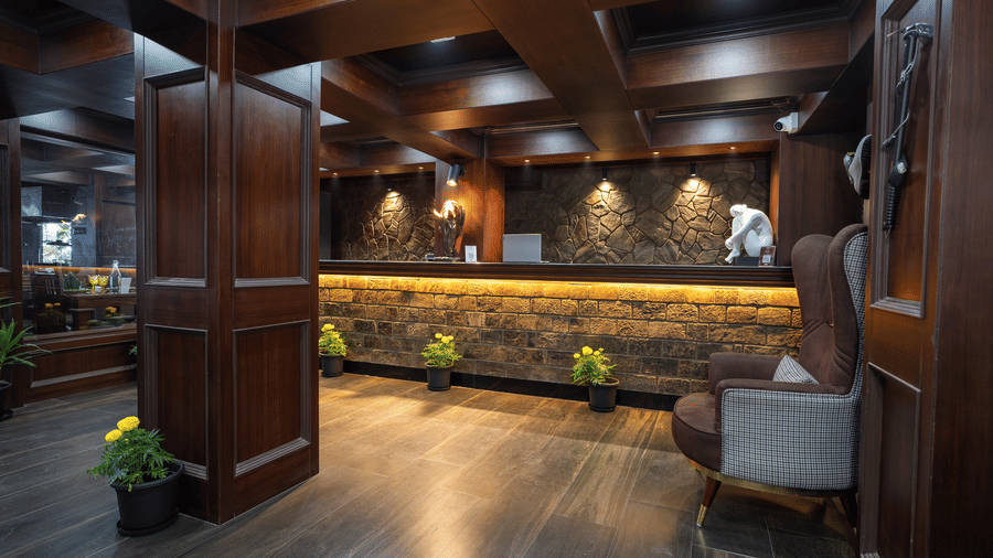 Reception desk and lobby lounge area inside The West Gate Posada By Summit, Darjeeling, featuring wooden panelling, flooring, and armchairs.