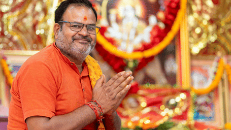 A man in an orange shirt with his hands in a prayer position smiling in front of a brightly decorated shrine.