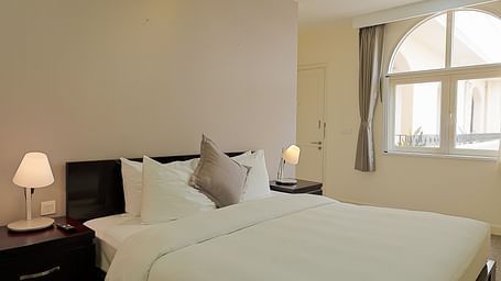 A room with a white bed, bedside tables, lamps, and a large window | Four-bedroom Suite | Central Mansions, Phnom Penh