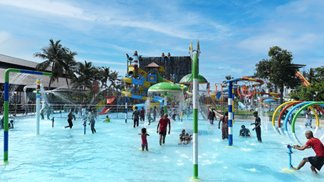 Children and adults in a shallow splash pool with water jets, spray arches and a mushroom fountain, with slides in the background at MGM Dizzee World, Chennai.