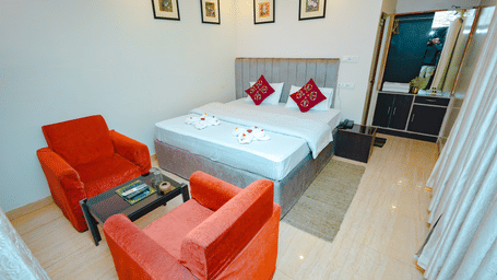 A spacious hotel bedroom featuring a double bed, white bedding with red accents, and a seating area with two orange armchairs.