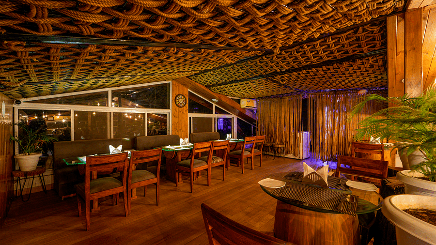 A shot taken from an angle of Ruttba which has wooden interiors and chairs and tables with bright lighting - DLS Whispering Winds
