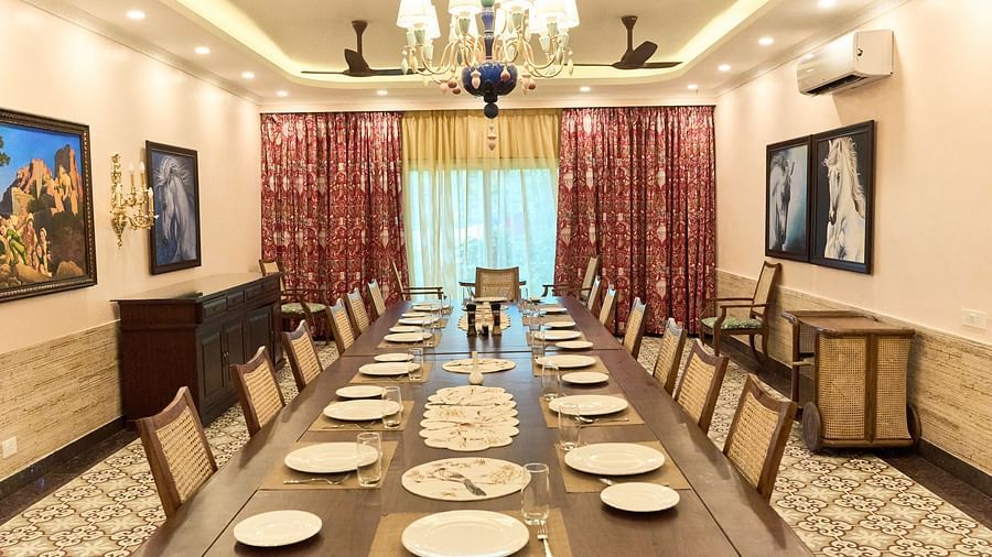 At Mayfair Manor, Alibag, a spacious dining table is beautifully arranged for a delightful culinary experience, enhanced by wall-mounted paintings, an elegant chandelier above, and expansive windows with curtains tastefully drawn to the side