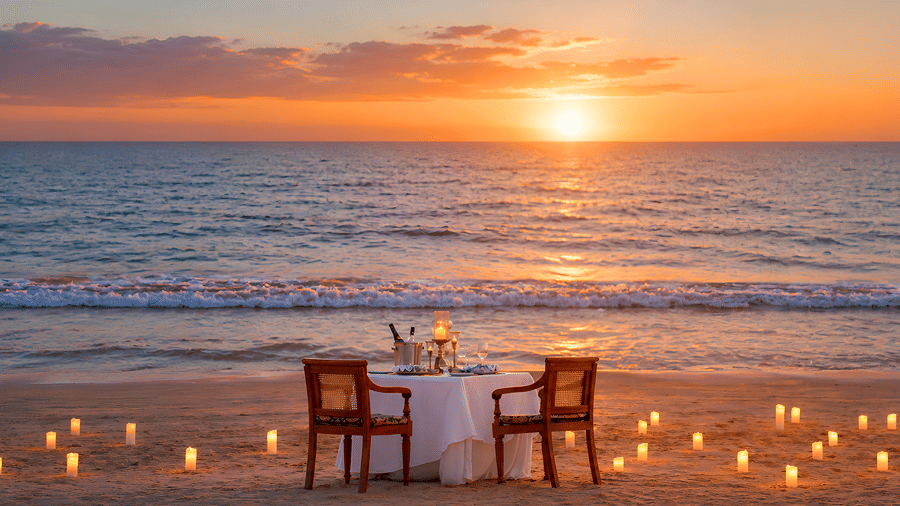Romantic dining setup for two on a sandy beach surrounded by candles during sunset at MAYFAIR Palm Beach Resort, Gopalpur-on-Sea.