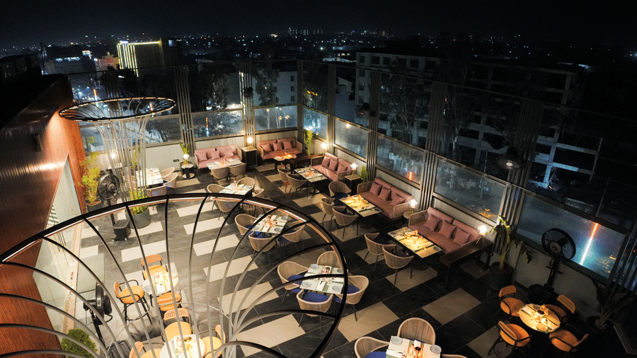 Rooftop dining area at Ramada Encore by Wyndham Chandigarh Zirakpur, featuring modern outdoor seating with cushioned sofas and dining tables, warm ambient lighting, glass railings, and a panoramic night view of the city skyline.